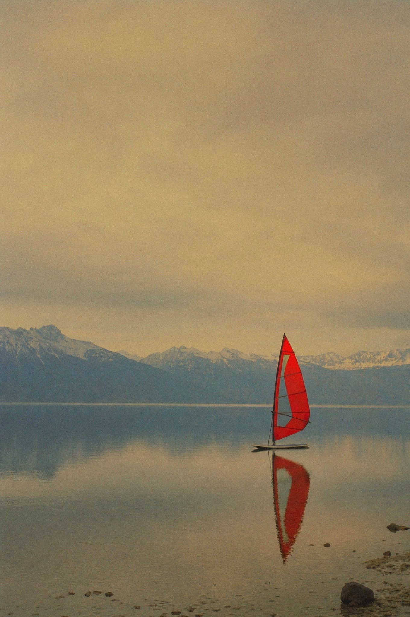 Archive grade — a single windsurf board with a red sail on the still water of lake Chiemsee, the Alps behind