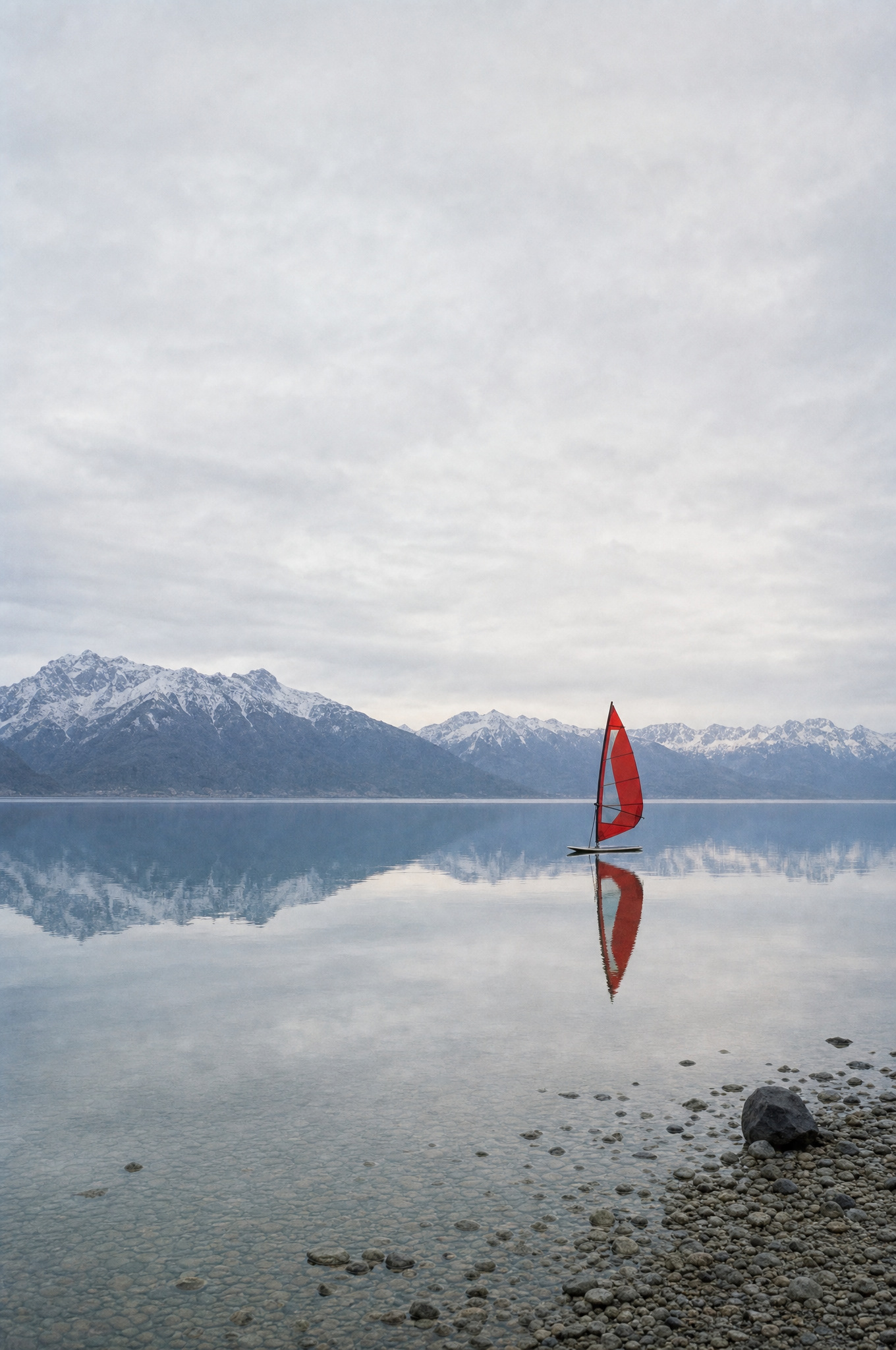 Campaign — windsurf board with a poppy-red sail resting on a still alpine lake at dawn, snow-dusted mountains behind